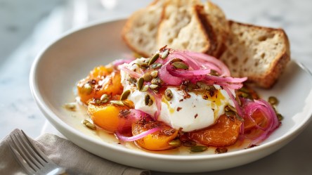 Ein Teller mit gebackenen Aprikosen und Burrata, die mit gerösteten Kürbiskernen und eingelegten roten Zwiebeln garniert sind. Das Ganze ist mit frischem knusprigem Brot serviert. Neben dem Teller liegt eine Gabel.