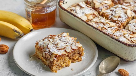 Ein Teller mit einem Stück der gebackenen Mandelcroissant-Haferflocken. Daneben steht die Auflaufform voll mit den Baked Oats. Außerdem sind Bananen, einzelne Mandeln, ein Löffel und ein Glas Honig im Bild zu sehen.