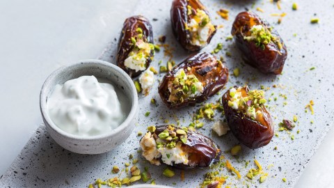Ein grauer Steinteller, auf dem einige gefüllte Datteln liegen, die zuvor gegrillte Dattel-Feta-Spieße waren. Die Datteln werden mit einer kleinen Schale Joghurt-Dip serviert.