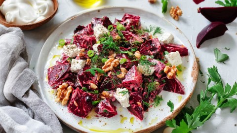 Ein großer Teller mit einer Portion griechischem Rote-Bete-Salat. Links neben dem Teller liegt ein hellgraues Küchenhandtuch. Außerdem sind eine Schüssel Joghurt sowie Stücke von Roter Bete, frische Petersilie und Walnüsse zu erkennen, die alle um den Teller herum liegen.