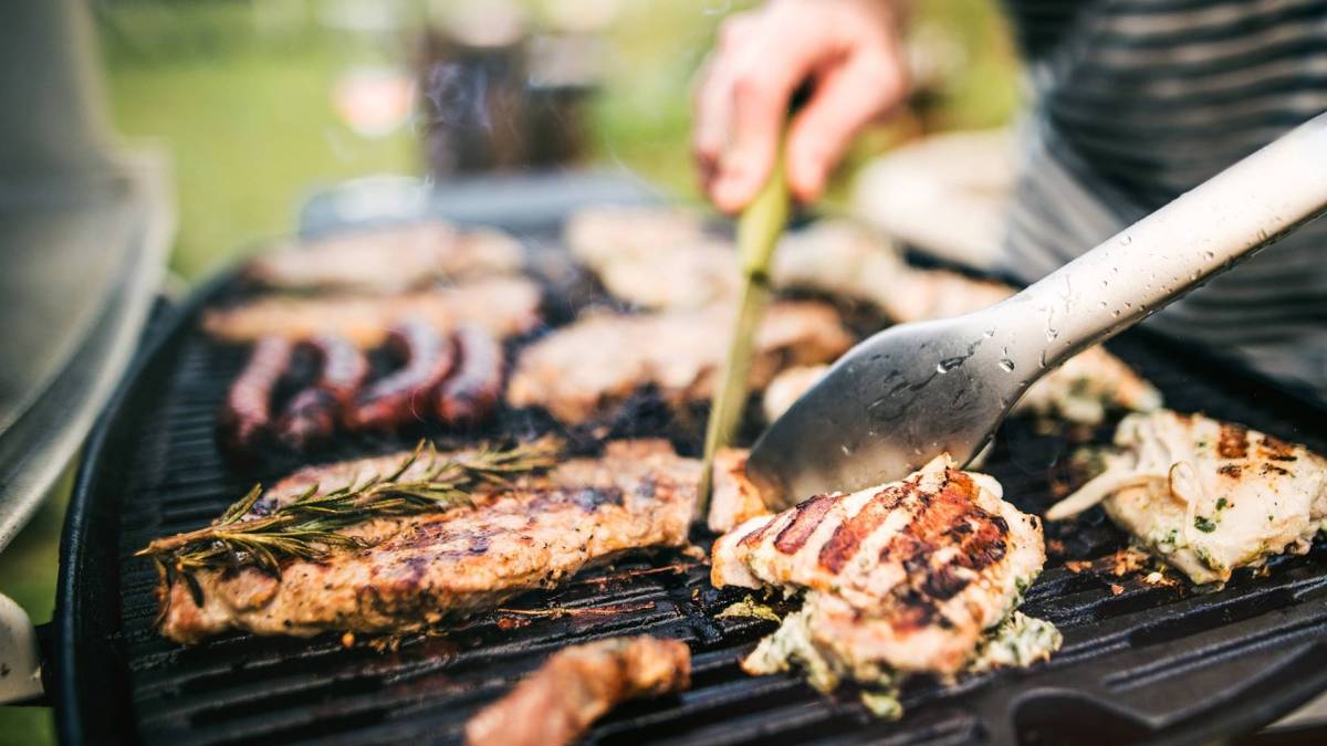Ein Grill, auf dem einiges an Grillfleisch liegt. Man kann angedeutet jemand erkennen, der das Grillfleisch mit Grillwerkzeug wendet.