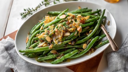 Ein ovalförmiger Teller mit einer großen Portion grüner Bohnen mit brauner Butter und gerösteten Mandeln. Der Teller steht auf einem dunklen Holzbrett. Außerdem ist ein graues Küchenhandtuch drapiert und man kann im Hintergrund einen Thymianzweig erkennen.