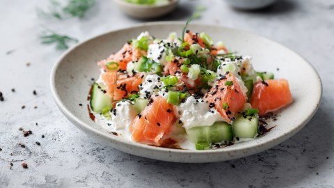 Ein Teller mit Gurkensalat mit Hüttenkäse und Lachs. Das Ganze ist mit schwarzem Sesam, Frühlingszwiebeln und Dill garniert.