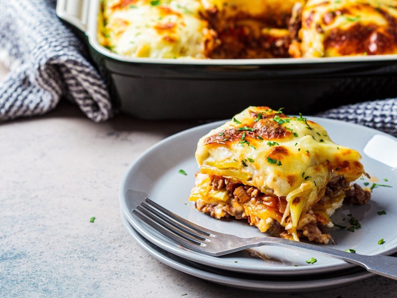 Ein Stück Gyros-Kartoffel-Auflauf liegt vor einer Auflaufform mit noch mehr Auflauf darin.