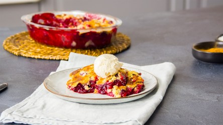 Ein Stück Himbeerkuchen-Auflauf mit einer Kugel Vanilleeis auf einem Teller. Im Hintergrund steht eine Auflaufform mit dem Auflauf.
