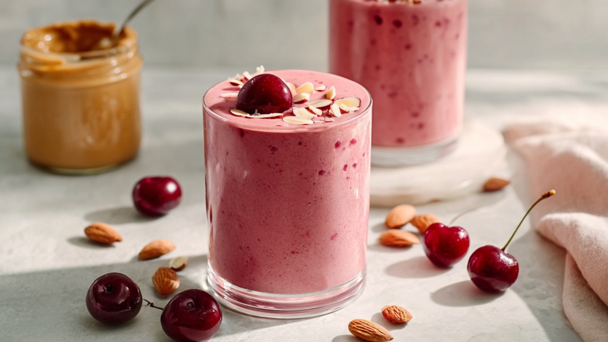 Ein Glas Kirsch-Mandel-Smoothie, das mit einer frischen Kirsche und einigen Mandelsplittern garniert ist. Ringsum liegen braune Mandeln und einige Kirschen verteilt. Im Hintergrund kann man ein weiteres Glas mit Smoothie erkennen sowie ein Glas mit Mandelmus, in dem ein Löffel steckt.