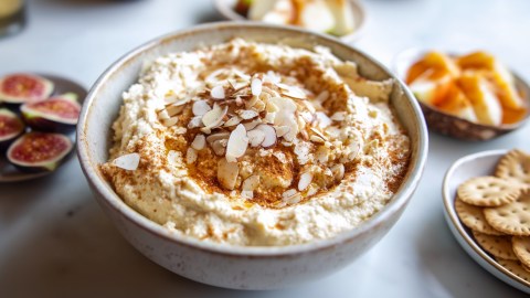 Eine Schüssel mit süßem Kokos-Hummus, das mit gerösteten gehackten Mandeln und Zimt garniert ist. Um die Schüssel mit dem Kokos-Hummus herum sind kleinere Teller mit Crackern, Feigen und Apfelschnitzen zu sehen.