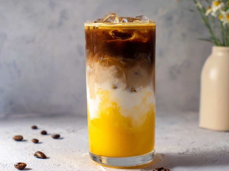 Ein großes Glas mit Mango Iced Latte, das vor einem hellen minimalistischen Hintergrund platziert ist. Es liegen ein paar Kaffeebohnen verstreut. Im Hintergrund steht eine beige Vase mit Kamillen.
