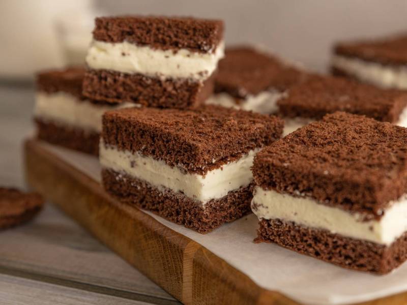 Ein Holzbrett, das mit Backpapier belegt ist und auf dem einige quadratische Stücke Milchschnitte-Kuchen liegen.