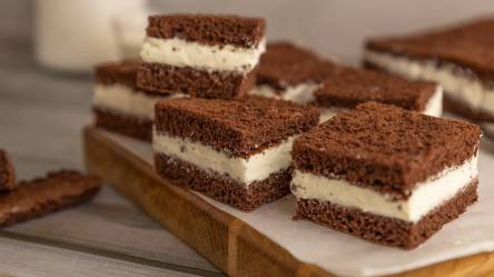 Ein Holzbrett, das mit Backpapier belegt ist und auf dem einige quadratische Stücke Milchschnitte-Kuchen liegen.