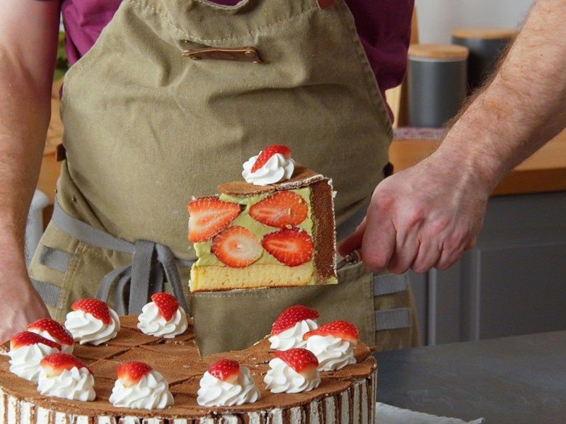 Ein Stück Milchschnittenkuchen wird aus dem Kuchen gehoben. Im Hintergrund sieht man ein Küchenset.
