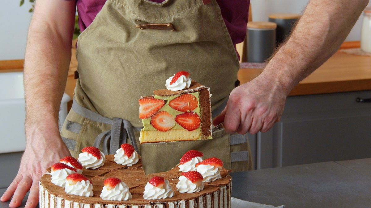 Ein Stück Milchschnittenkuchen wird aus dem Kuchen gehoben. Im Hintergrund sieht man ein Küchenset.