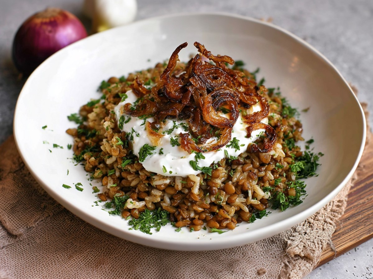Ein Teller mit einer großen Portion Mujaddara, also Reis mit Linsen und karamellisiertem Zwiebeln. Alles ist mit etwas frischem Joghurt und frischen gehackten Kräutern. Im Hintergrund liegt ein rote Zwiebel und eine Knoblauchknolle.