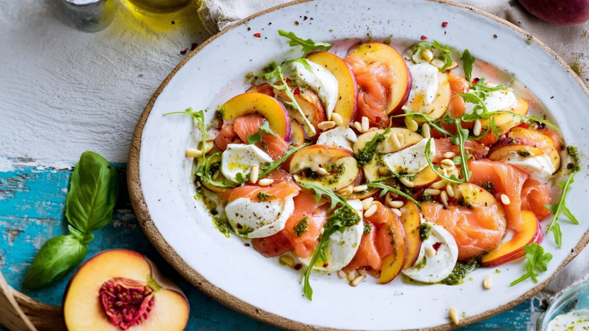Ein großer Servierteller mit Nektarinen-Räucherlachs-Caprese. Alles ist mit Pinienkernen, frischem Rucola und etwas Basilikum-Pesto angerichtet. Um den Teller herum liegen eine halbe Nektarine, etwas frischer Basilikum und man kann angeschnitten Essig- und Ölgefäße erkennen.