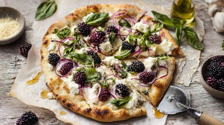 Eine Pizza mit Brombeeren und Ricotta, die mit frischem Basilikum garniert und zusätzlich mit roten Zwiebeln belegt ist. Die Pizza liegt auf Backpapier und ist mit Honig beträufelt. Daneben stehen eine Schale mit Brombeeren und eine mit geriebenem Käse. Außerdem ist ein Pizzaschneider zu erkennen.
