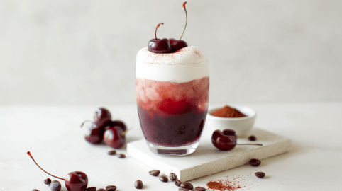 Ein Glas mit Red Velvet Kaffee auf einem kleinen Brett. Das Glas ist umgeben von einigen Kirschen und Kaffeebohnen.