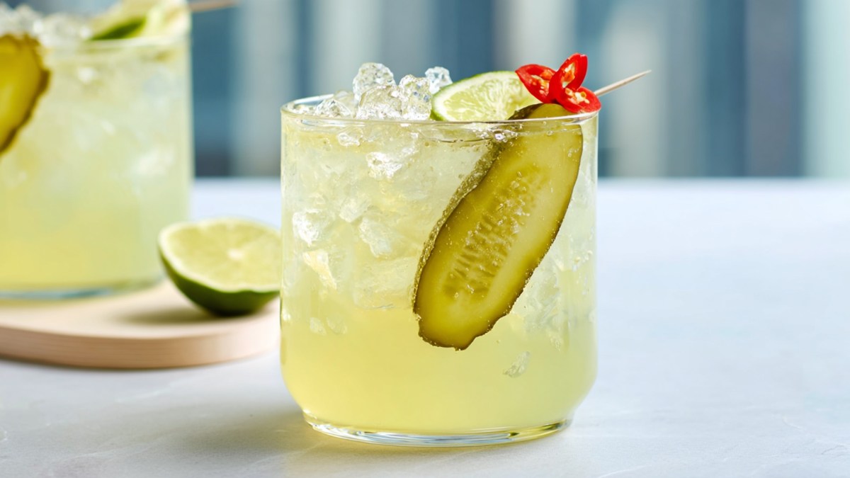 Ein Glas mit scharfer Essiggurken-Limonade, die mit einer langen Scheibe Essiggurke, einem Limettenstück und feinen Chiliringen garniert ist. Im Hintergrund ist ein weiteres Glas zu sehen, das auf einem Brett steht, auf dem auch eine angeschnittene Limette liegt.