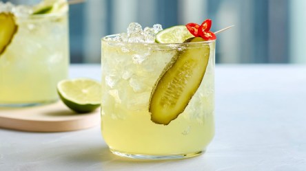 Ein Glas mit scharfer Essiggurken-Limonade, die mit einer langen Scheibe Essiggurke, einem Limettenstück und feinen Chiliringen garniert ist. Im Hintergrund ist ein weiteres Glas zu sehen, das auf einem Brett steht, auf dem auch eine angeschnittene Limette liegt.