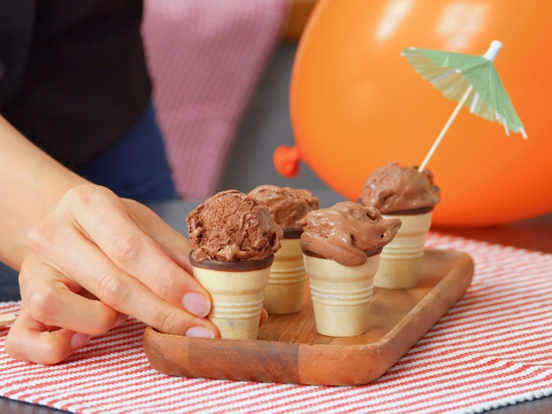Ein kleines Holzbrett, das auf einem rotgestreiften Tischuntersetzer steht. Auf dem Brett stehen einige Waffeln mit selbstgemachtem Schokoladen-Eis. In einer Eiskugel steckt ein kleines Cocktailschirmchen. Ein Hand greift nach einer der Eiswaffeln. Im Hintergrund kann man einen orangen Ballon erkennen.