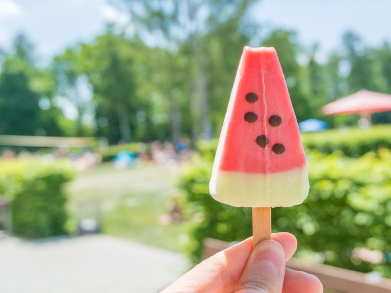 Snacks Freibad: Jemand hält im Freibad ein Eis am Stiel vor sich hoch.