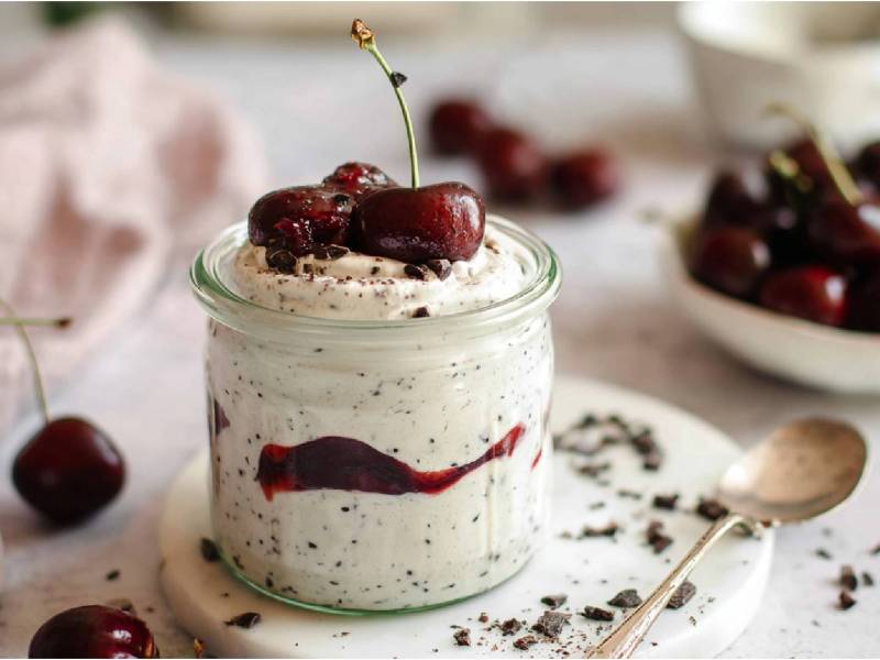Ein Glas mit Stracciatella-Quark mit Kirschkompott, Schale mit frischen Kirschen im Hintergrund