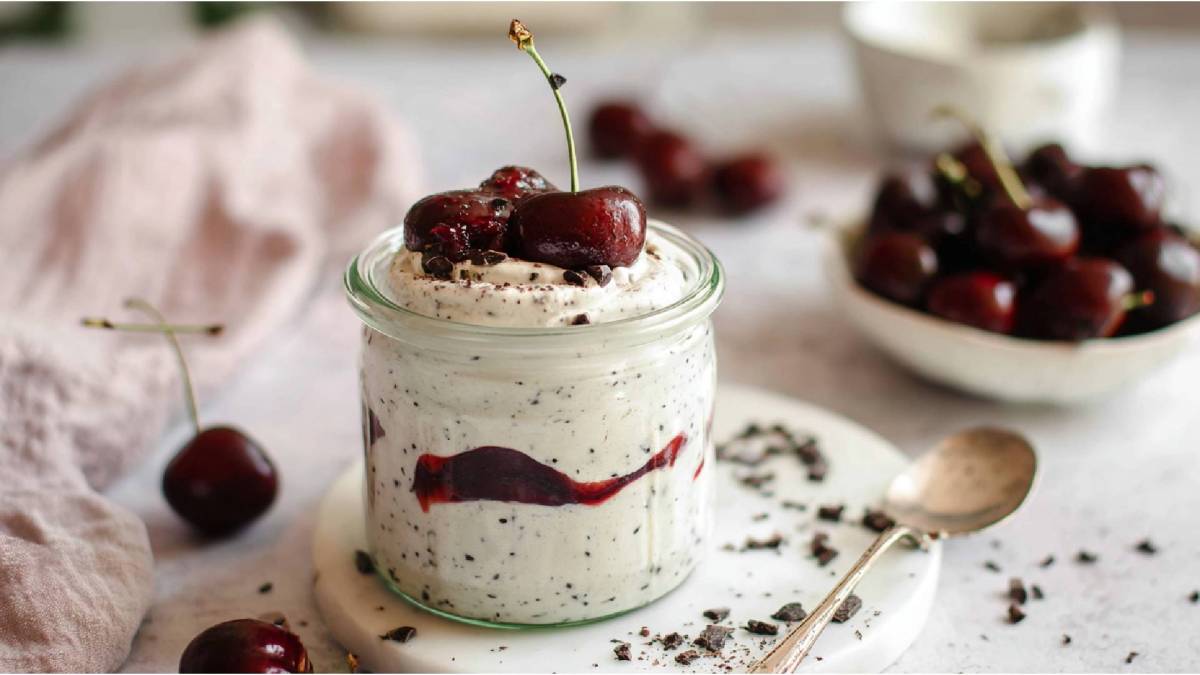 Ein Glas mit Stracciatella-Quark mit Kirschkompott, Schale mit frischen Kirschen im Hintergrund