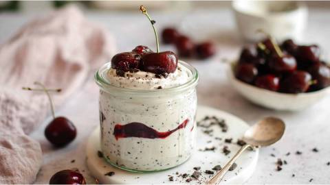 Ein Glas mit Stracciatella-Quark mit Kirschkompott, Schale mit frischen Kirschen im Hintergrund