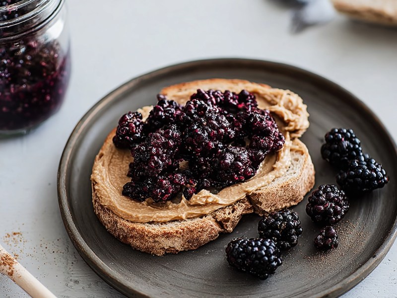 Ein Toast mit Mandelmus und Brombeeren, der auf einem dunklen rustikalen Teller liegt. Auf dem Teller liegen noch einige einzelne Beeren. Neben dem Teller liegt ein Holzlöffel mit Zimt. Im Hintergrund kann man ein Glasgefäß mit frischen Brombeeren sehen.