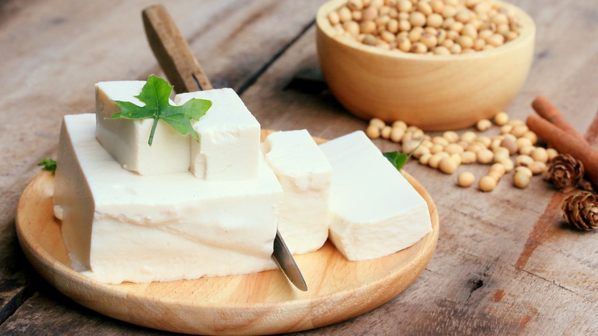 Ein Block Tofu und mehrere Stücke auf einem Holzbrett. Dahinter eine Schale mit Sojabohnen.