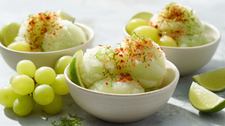 Drei Schalen Traubensorbet auf grauem Untergrund.c Sie sind mit Weintrauben und Limettenspalten angerichtet. Links liegen noch einige frische Trauben zwischen den Schalen.