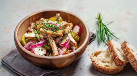 Eine Holzschüssel mit veganem Wurstsalat aus Räuchertofu. Neben der Schüssel liegen drei frische Scheiben Sauerteigbrot und ein kleiner Bund Schnittlauch.