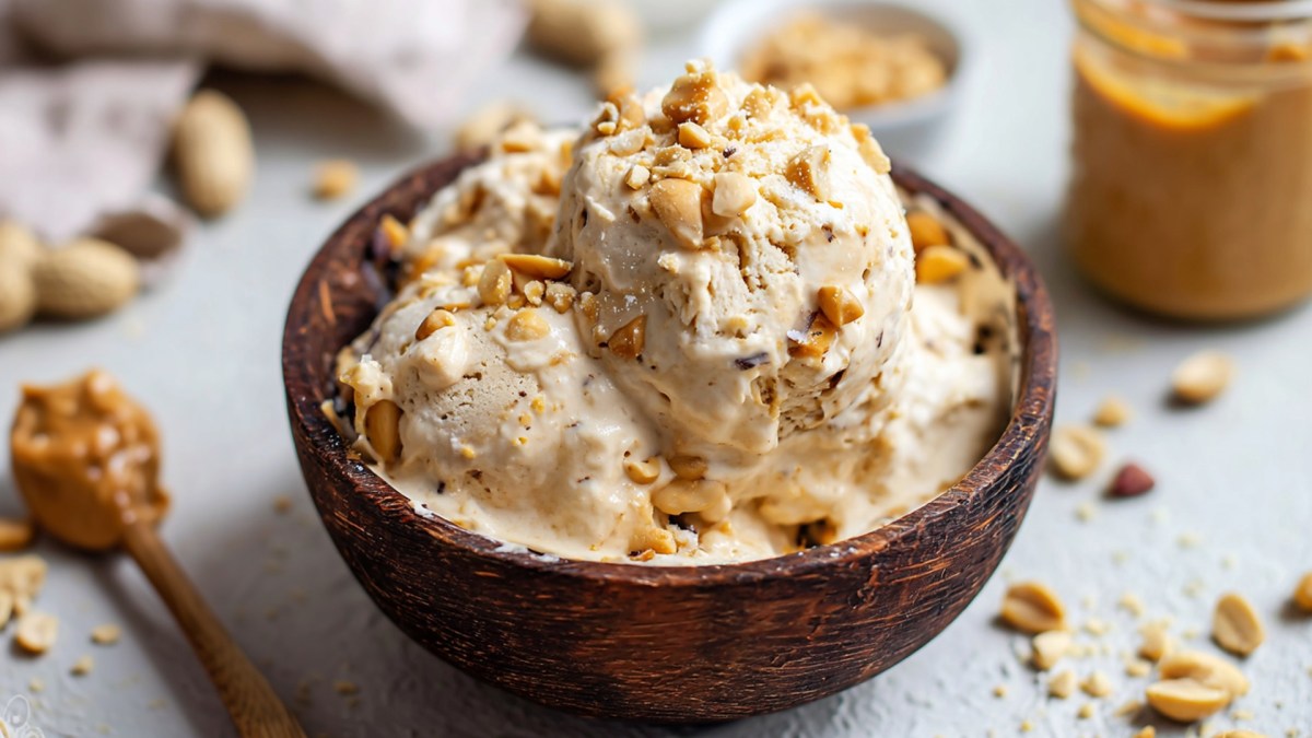 Eine kleine Holzschüssel, die mit einigen Kugeln veganem Erdnussbutter-Eis aus Seidentofu gefüllt ist und mit gehackten Erdnüssen garniert ist. Daneben liegt ein Löffel mit Erdnussbutter. Im Hintergrund steht ein Glas mit Erdnussbutter. Es liegen gehackte Erdnüsse und ganze Erdnüsse in der Schale herum.