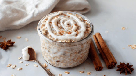Ein Glas Zimtschnecken Overnight Oats steht auf einer hellen Unterlage. Rechts neben dem Glas liegen zwei Zimtstangen sowie ein Sternanis. Links liegt ein Löffel mit dem Topping sowie ein weiterer Sternanis.