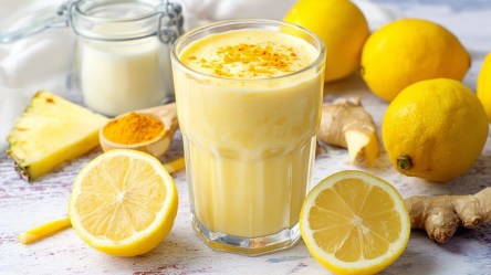 Ein Glas Zitronen-Smoothie. Ringsum liegen ganz und aufgeschnittene Zitronen, eine Kurkumawurzel sowie ein Löffel mit Kurkumapulver und ein Glasgefäß mit Milch darin.