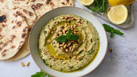 Eine Schale mit Zucchini Baba Ganoush, das mit gerösteten Pinienkernen, etwas Olivenöl und frischer Minze garniert ist. Ringsum liegt Fladenbrot, frische Minze und Zitronen verteilt.
