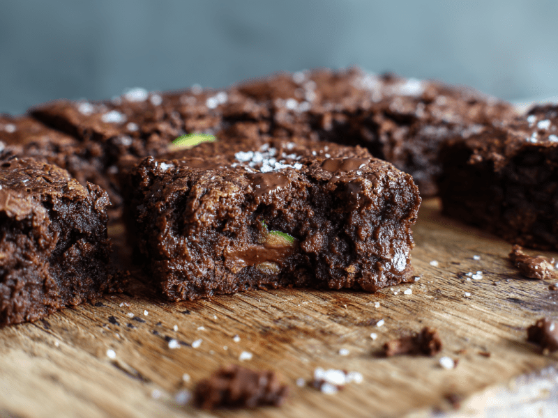Mehrere Zucchini Brownies liegen auf einem Holzbrett. Sie sind angeschnitten und geben den Blick auf das Innere frei.