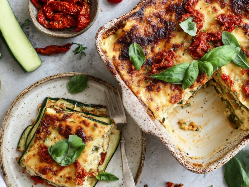 Draufsicht auf eine Auflaufform mit Zucchini-Feta-Lasagne. Ein Stück davon liegt auf einem Teller daneben. Ringsum sind frische Zutaten verteilt wie Zucchini, eine Schale mit getrockneten Tomaten, eine Glasgefäß mit Olivenöl, frisches Basilikum.