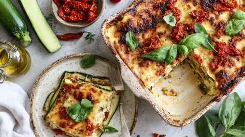 Draufsicht auf eine Auflaufform mit Zucchini-Feta-Lasagne. Ein Stück davon liegt auf einem Teller daneben. Ringsum sind frische Zutaten verteilt wie Zucchini, eine Schale mit getrockneten Tomaten, eine Glasgefäß mit Olivenöl, frisches Basilikum.