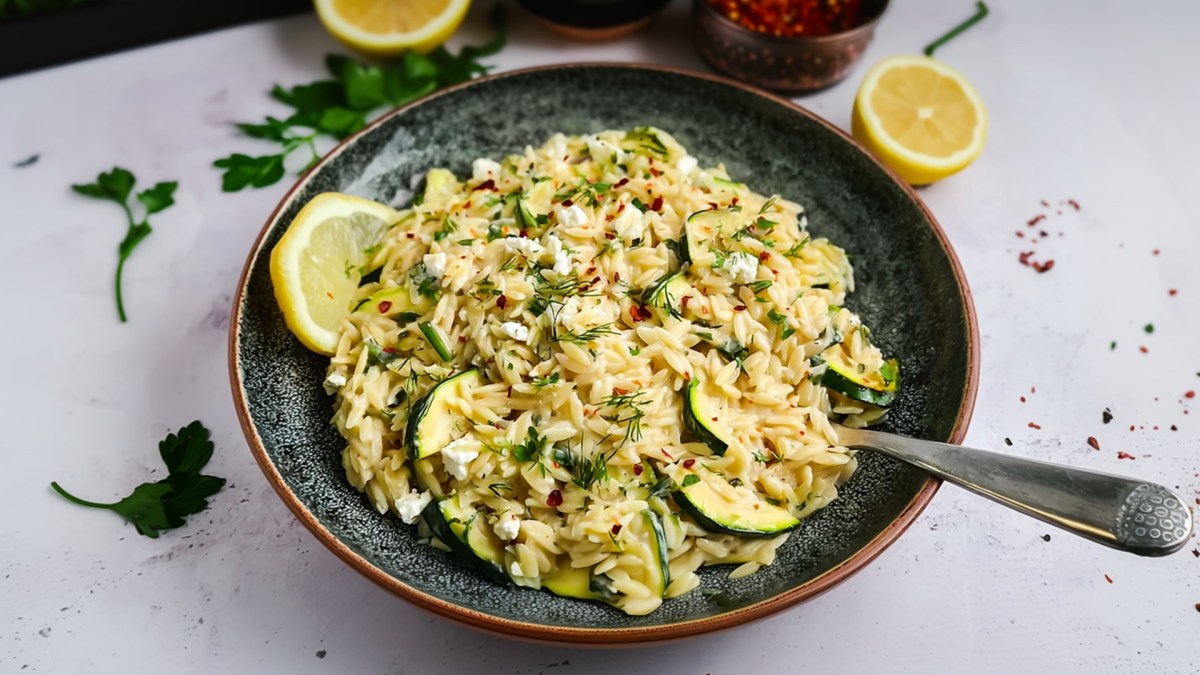Ein großer tiefer Teller mit Zucchini-Feta-Orzo, der mit frischer Petersilie und Chiliflocken garniert ist. Außerdem liegt eine Zitronenscheibe dabei. Um den Teller herum sind Zutaten zu sehen wie halbe Zitronen, frische Petersilie und eine Schale mit Chiliflocken.