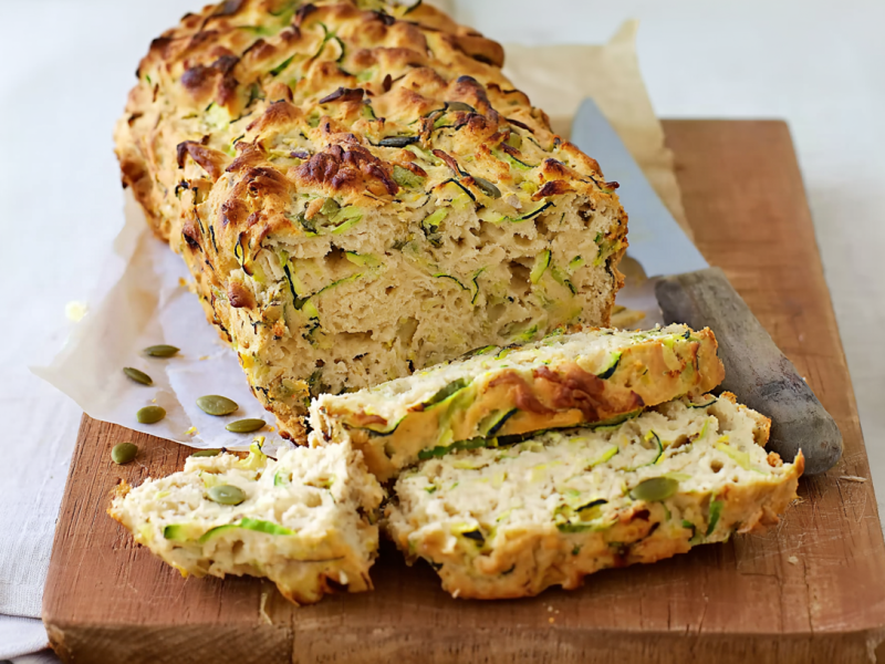 Ein angeschnittenes Zucchini-Käsebrot auf einem Holzbrett. Rechts neben dem Brot liegt ein Messer. Rechts neben dem Brett sieht man eine Schale mit Kürbiskernen.