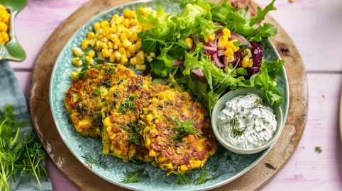 Zucchini-Mais-Puffer, serviert auf einem Teller mit Kräuterdip, Salat und frischem Mais.