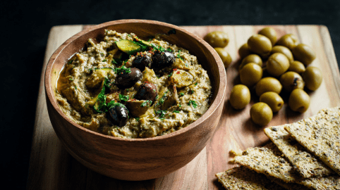 Eine Holzschale mit Zucchini-Oliven-Aufstrich, daneben liegen einige grüne Oliven.