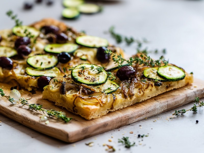Eine Zucchini-Pissaladière, mit Zucchini, karamellisierten Zwiebeln, schwarzen Oliven und Sardellen. Die Pissaladière liegt auf einem Holzbrett. Ringsum liegt frischer Thymian verstreut.