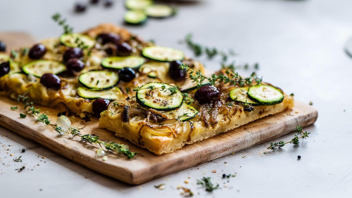 Eine Zucchini-Pissaladière, mit Zucchini, karamellisierten Zwiebeln, schwarzen Oliven und Sardellen. Die Pissaladière liegt auf einem Holzbrett. Ringsum liegt frischer Thymian verstreut.