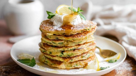 Ein Stapel Zucchini-Zitronen-Pancakes mit Ricotta, die mit einem Klecks Ricotta, einer Zitronenscheibe und frischer Minze garniert sind. Von oben wird Honig drüber geträufelt. Im Hintergrund ist ein weißes Laken drapiert und eine weiße Vase ist zu erkennen.