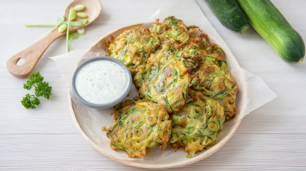 Zucchinipuffer auf einem Teller. Daneben ein Dip und eine Holzgabel.