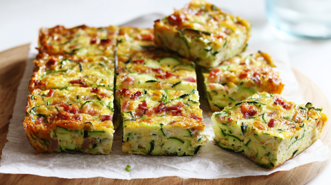 Zucchinischnitten mit Ei und Bacon auf einem Stück Backpapier, das auf einem Holzbrett liegt.