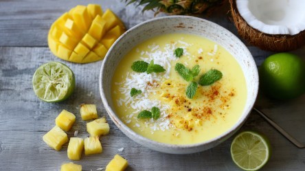 Eine Ananas-Kaltschale, die umringt ist von einer gewürfelten und aufgefächerten Mango, Ananasstückchen, einer ausgepressten und einer normalen Limettenhälfte und einer ganzen Limette und einer halben Kokosnuss.