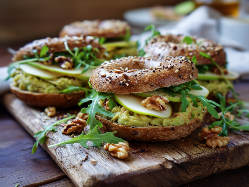 Vier Avocado-Birnen-Bagel liegen auf einem rustikalen Holzbrett. Sie sind mit Rucola und Walnüssen verfeinert. Im Hintergrund sieht man ein graues Küchentuch und eine halbierte Avocado