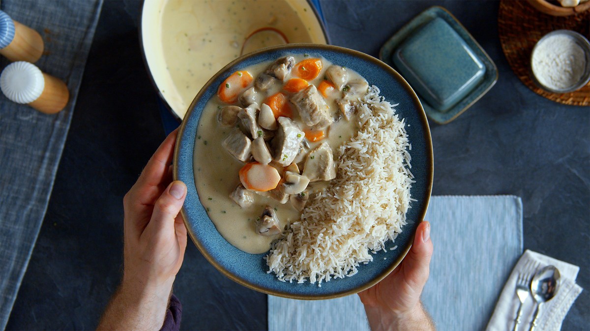 Ein dunkelblauer Teller mit einer Portion Blanquette de Veau mit Reis. Der Teller wir von zwei Händen gehalten. Draufsicht. Unter dem Teller kann man den Kochtopf mit dem Blanquette de Veau erkennen, sowie einen hellblauen Tischläufer, Salz und Pfefferstreuer und Besteck sowie sonstige Küchenutensilien.
